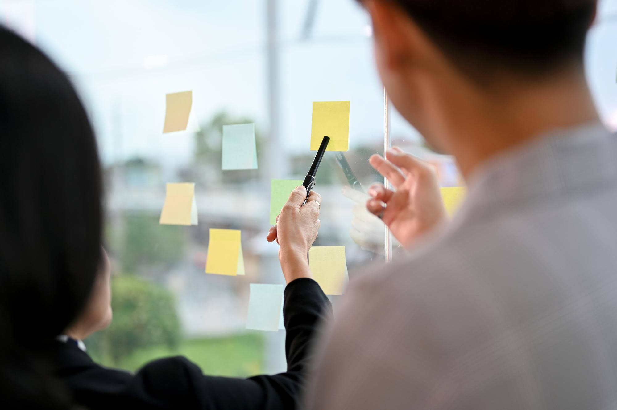 Kaksi henkilöä katselee lasiseinää, jossa on kiinnitettynä muistilappuja. Nainen osoittaa lappua mustalla kynällä. Valoisa ja avoin tunnelma.